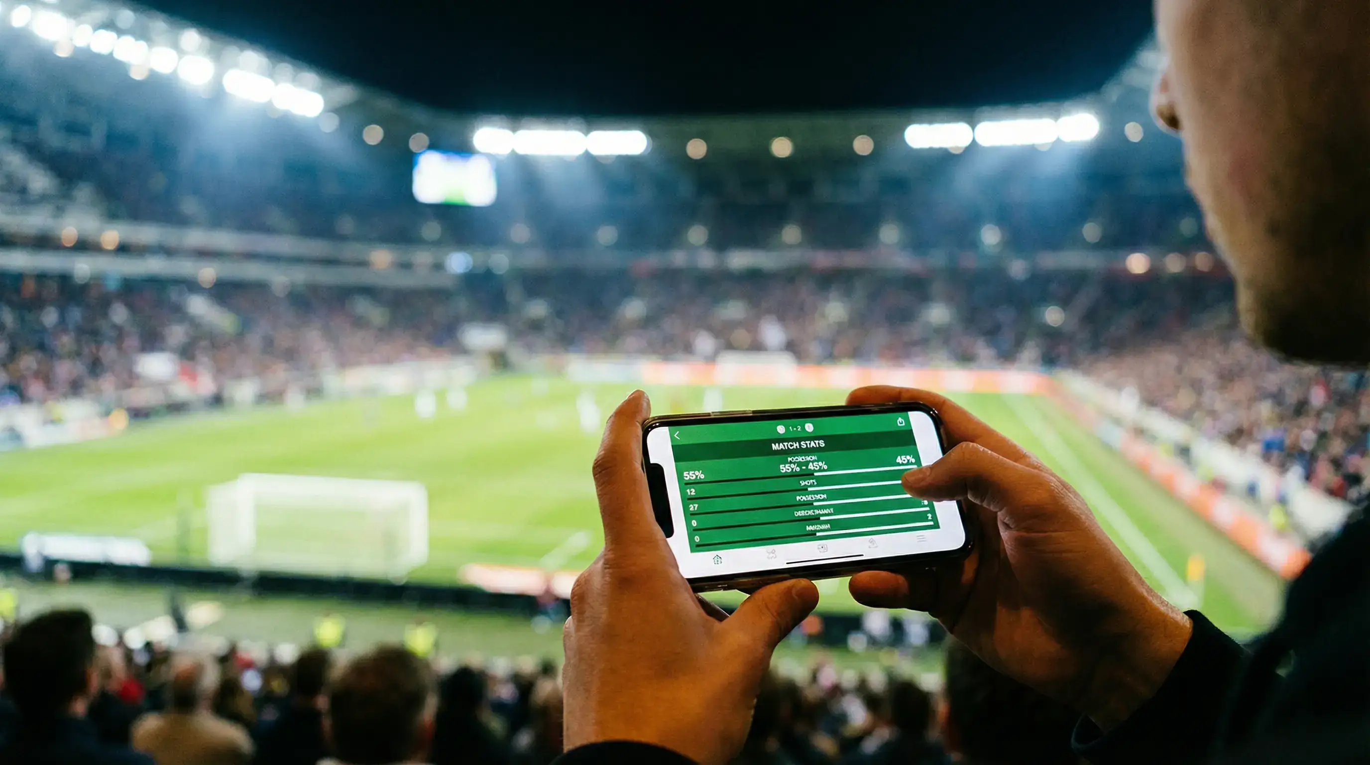 Hænder der holder en smartphone foran et oplyst fodboldstadion om aftenen med grøn bane i baggrunden